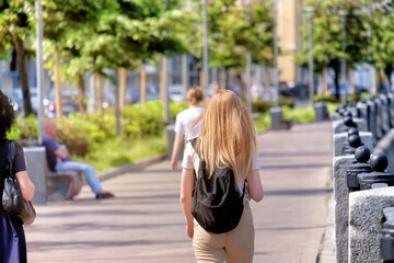 young female walking along city street on summer day against blurred urban background. Authentic lifestyle scene of gold hair woman in sand colored pants and white t-shirt with black backpack on back