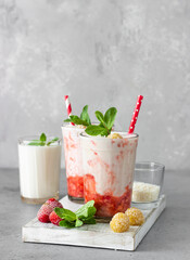 Layered strawberry and raspberry smoothie or milkshake in glasses decorated with mint, raspberry and coconut flakes on grey table. Healthy food for breakfast or snack.
