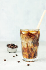 Iced coffee with milk in a tall glass with roasted coffee beans on light grey concrete background. Summer refreshment drink. Selective focus.