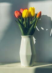 Bouquet of red and yellow tulips stands in a white vase on a white background. Flowers in a vase. mother's day. Red and yellow tulips in a white geometric ceramic vase stand. fresh flowers in a vase