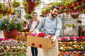 Two smiling florists holding boxes with orders and walking trough greenhouse. In foreground is man.