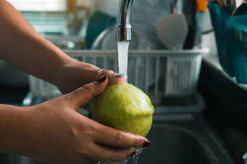 Woman hand is holding a guava and washing with water at the tap in the kitchen at home.