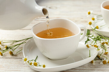 Pouring tasty chamomile tea into cup on white wooden table