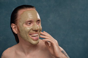 Happy laughing man with wet long hair in green clay mask on his face waiting of spa procedures.