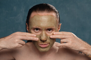 A man with long wet hair is applying a green clay mask with his hands.