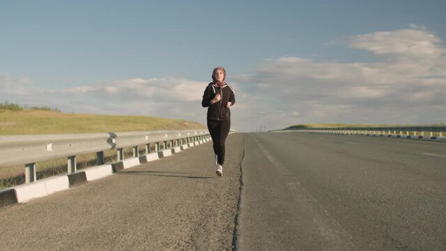 An Eastern-looking girl athlete with a scarf on her head runs along the side of an empty highway against the blue sky. A girl in a tracksuit of Eastern appearance is running on an empty track