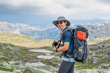 Young Asian man traveller with backpack taking picture while trekking in Trolltunga mountain cliff,...