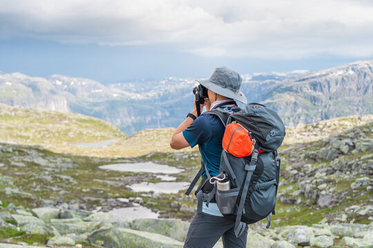 Young Asian man traveller with backpack taking picture of mountains landscape while trekking in Trolltunga mountain cliff, famous trekking route in Odda, Norway, Scandinavia