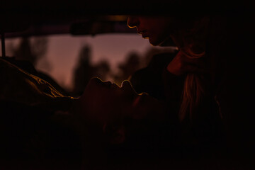 Silhouette of lovers in the car in the back seat. Loving couple retired at dusk to spend the evening together. The girl bent to kiss the guy who is lying below and looking into her eyes. Passion 