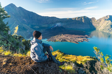 Young Asian man trekker sitting on Sembalun crater in Rinjani active volcano mountain, Lombok island in Indonesia