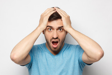 Shock content. Surprised young man in blue t-shirt shouting, holding his head with both hands, isolated on gray background