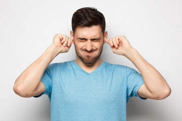 Young man in blue tshirt closing ears suffering from loud noise, isolated on gray background