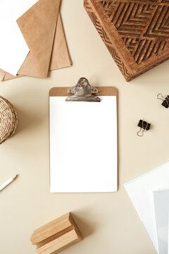 Clipboard tablet pad with blank paper sheet on beige table. Artist home office desk workspace with wooden casket, pencil, envelopes and stationery. Flat lay, top view mockup with empty copy space