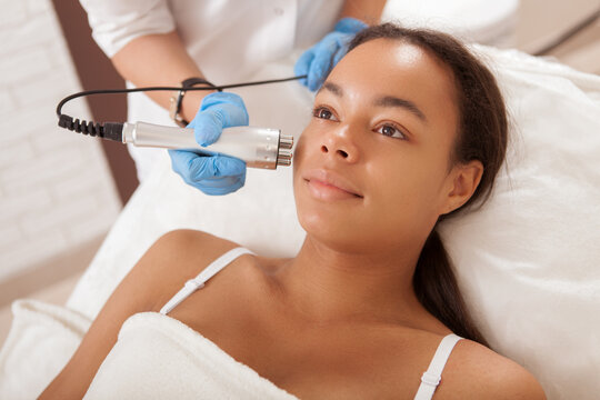 Happy beautiful African woman smiling while getting skin tightening treatment by beautician