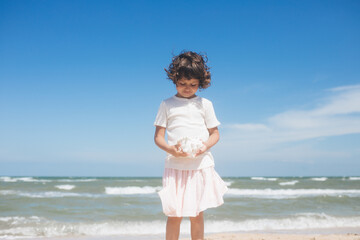 Fototapeta na wymiar Little girl wearing light t-short and light pink skirt at the sea beach with a big beautiful seashell. Summer vacation