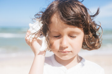 Little girl wearing light t-short and light pink skirt at the sea beach with a big beautiful seashell. Summer vacation