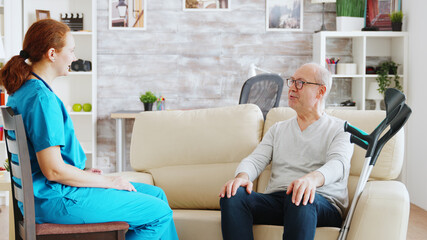 Fototapeta premium Elderly disabled man with crutches next to him talking with a nurse in bright and cozy nursing home
