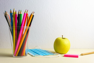 Farbstifte in einem Plastikbeh&auml;lter, ein blauer Mundschutz und ein gr&uuml;ner Apfel vor einem wei&szlig;en Hintergrund