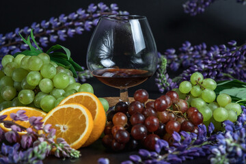 A glass of strong alcohol on a dark background. Still life of fruit and flowers. Grapes, oranges, and lupins. Ice cube. Cognac glass.
