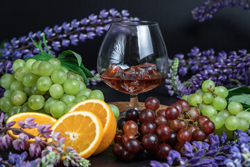 A glass of strong alcohol on a dark background. Still life of fruit and flowers. Grapes, oranges, and lupins. Ice cube. Cognac glass.