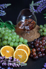 A glass of strong alcohol on a dark background. Still life of fruit and flowers. Grapes, oranges, and lupins. Ice cube. Cognac glass.