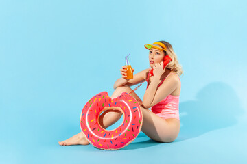 Full length, pensive woman in bathing suit sitting with rubber ring, drinking fresh juice or cocktail and talking on phone handset with serious attentive expression. indoor studio shot isolated