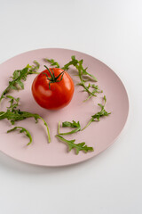 Fresh green arugula leaves and tomato on a pink plate on the left on a white table. Healthy diet. Vertical. Close-up