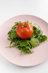 Salad cooked with fresh spinach and arugula leaves and tomato on a pink plate in the center on a white table. Healthy diet. Close-up. Vertical