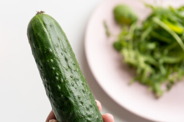 Fresh green cucumber in a female hand over a white table with a pink plate with spinach and arugula salad. Top view