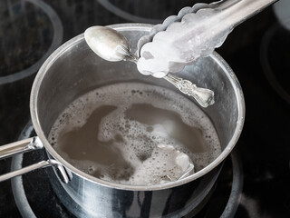 workshop for cleaning tarnished silver with aluminum foil and baking soda - tongs removes clean silver spoon from boiling solution of baking soda with aluminum foil in pot on electric stove