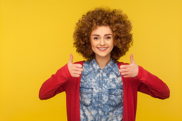 Like gesture! Happy successful woman with curly hair in casual style jeans shirt showing thumbs up, satisfied with excellent result, good feedback. indoor studio shot isolated on yellow background