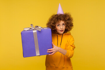 Portrait of amazed curly-haired woman holding huge gift box near ear and listening what's inside,...