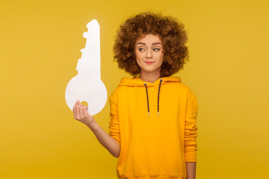 Portrait of funny curly-haired young woman in urban style hoodie looking at big paper key with interested curious expression, dreaming of own home. indoor studio shot isolated on yellow background