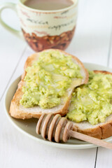 Tasty toasts with avocado and honey and a cup of coffee for breakfast on plate on white wooden table. Vertical. Close-up