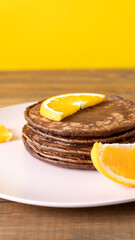 Delicious chocolate pancakes with slices of orange for breakfast on a pink plate on wooden table on yellow background. Fresh, sweet and healthy dessert. Vertical