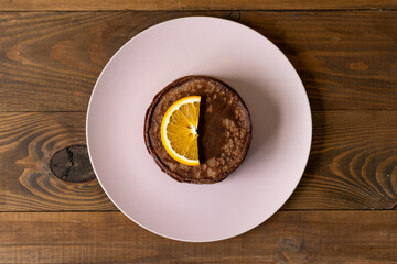 Delicious chocolate pancakes with slices of orange for breakfast on a pink plate on wooden background. Fresh, sweet and healthy dessert. Top view