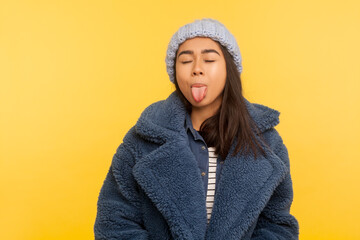 Portrait of carefree girl wearing warm winter hat and fur coat, stylish urban outfit, standing with closed eyes showing tongue out, glamour and fashion. studio shot isolated on yellow background