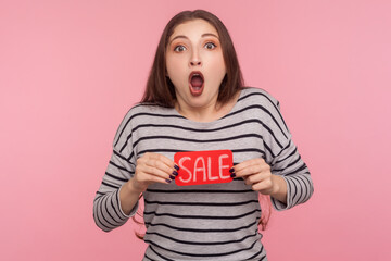 Wow, thrift shopping, discount! Portrait of surprised woman in striped sweatshirt holding Sale inscription and looking at camera with shocked expression. indoor studio shot isolated on pink background