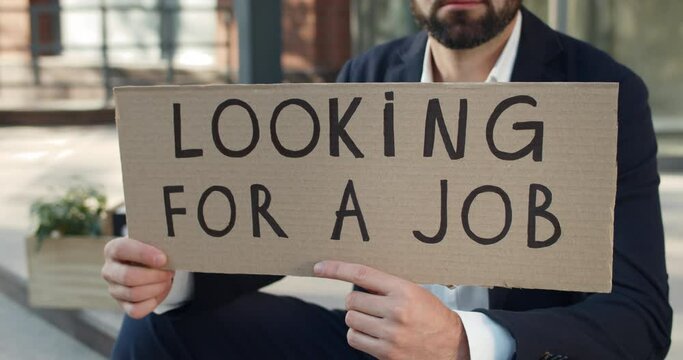 Crop view of male employee holding carton banner with looking for a job writing. Man searching for work while sitting at street. Concept of financial global crisis and unemployment