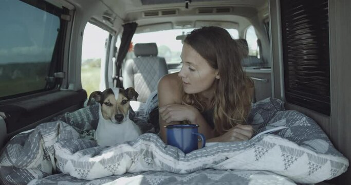 Young adult female on road trip inside Campervan reading book and playing with dog in bed