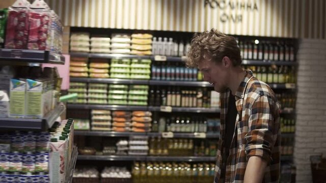 Young guy in supermarket. Caucasian handsome guy in plaid shirt choosing pasteurized milk from variety on the store shelves. Side view