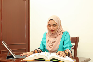 young Asian Malay Muslim woman wearing headscarf at home office student sitting at table talk mingle look at computer pone book document study discuss talk smile happy look at camera type