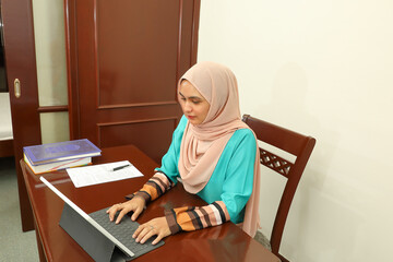 young Asian Malay muslim woman wearing headscarf at home office student sit at table computer book file paper study work