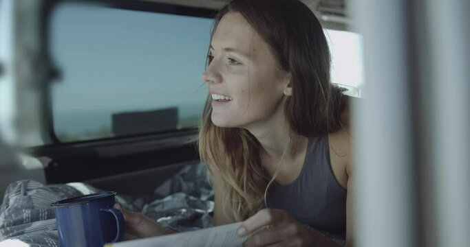 Young adult female on road trip inside Campervan reading book and drinking coffee smiling at boyfriend