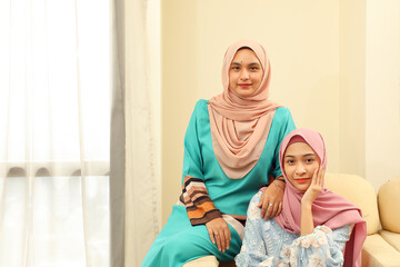 Two young Asian Malay Muslim woman wearing headscarf at home office student sitting on sofa look at camera smile happy