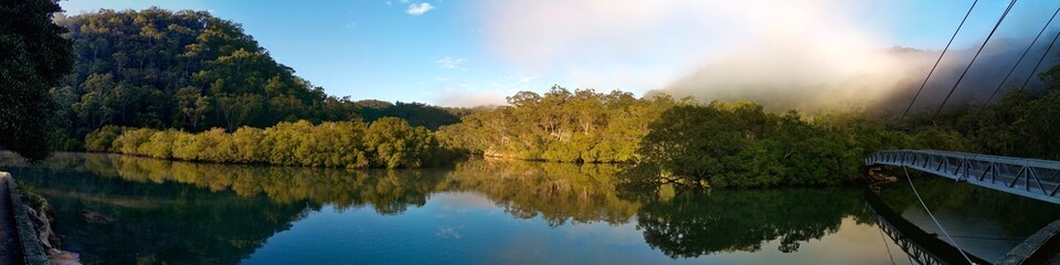 Fototapeta na wymiar Beautiful morning panoramic view of Cockle creek with reflections of blue sky, foggy mountains and trees, Mangrove boardwalk, Bobbin Head, Ku-ring-gai Chase National Park, New South Wales, Australia 