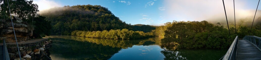 Beautiful morning panoramic view of Cockle creek with reflections of blue sky, foggy mountains and trees, Mangrove boardwalk, Bobbin Head, Ku-ring-gai Chase National Park, New South Wales, Australia

