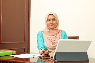 young Asian Malay Muslim woman wearing headscarf at home office student sitting at table talk mingle look at computer pone book document study discuss talk smile happy look at camera type