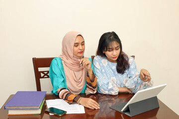Two young Asian Malay Muslim woman wearing headscarf at home office student sitting at table talk mingle look at computer book document study discuss read type write smile happy