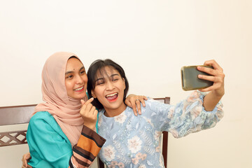 Two young Asian Malay Muslim woman wearing headscarf at home office student sitting at table phone computer book document selfie self portrait with smartphone care love